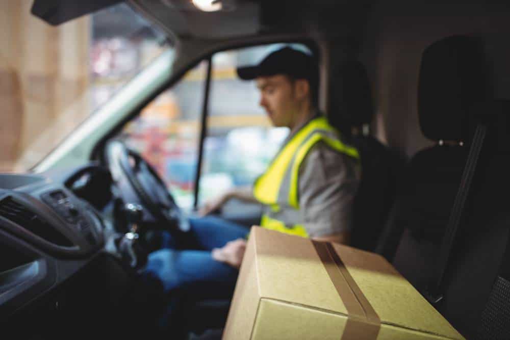 Delivery driver driving van with parcels on seat outside warehouse